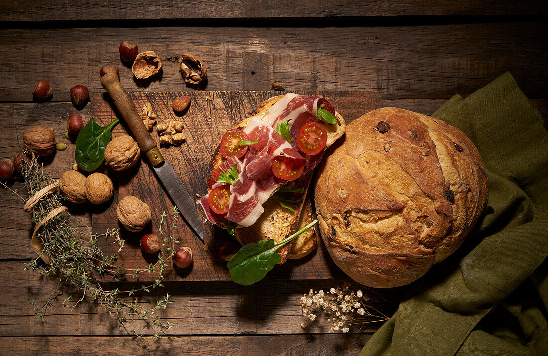 Draufsicht auf ein appetitliches hausgemachtes Sandwich aus frisch gebackenem Handwerksbrot mit Prosciutto- und Tomatenscheiben auf einem rustikalen Holztisch mit Nüssen und Kräutern
