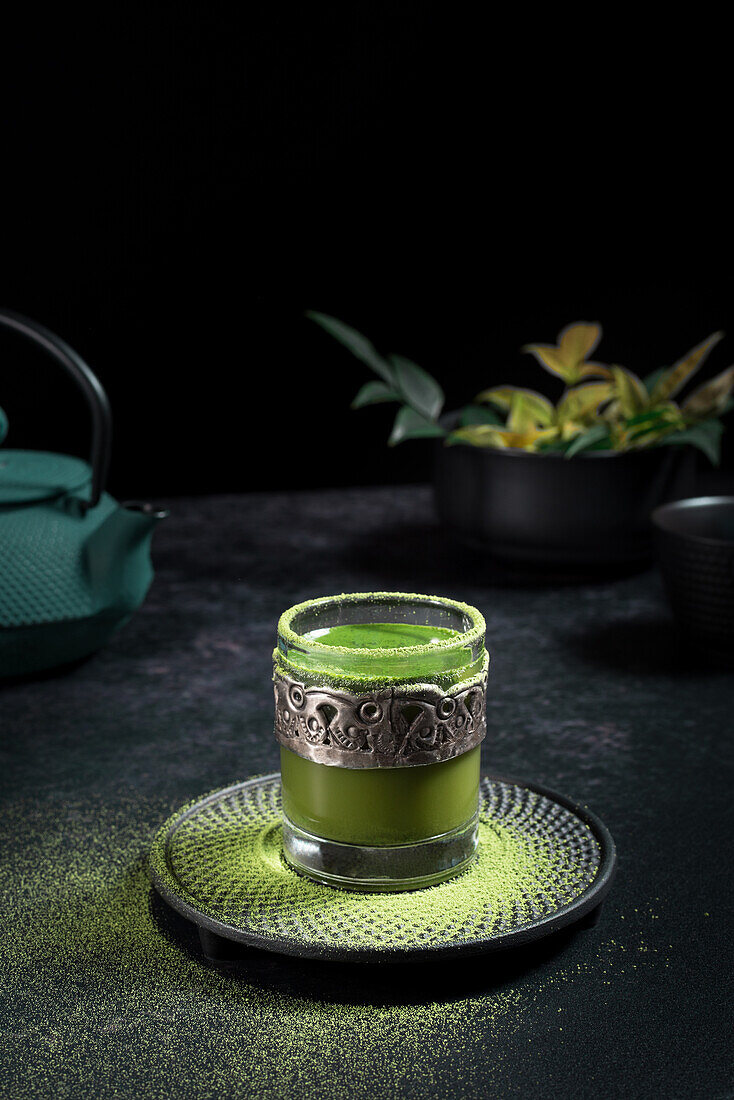 Stillleben Komposition mit traditionellem orientalischen Matcha-Tee in Glastasse mit Metallverzierung auf Tisch mit Keramikschalen und frischen grünen Blättern vor schwarzem Hintergrund