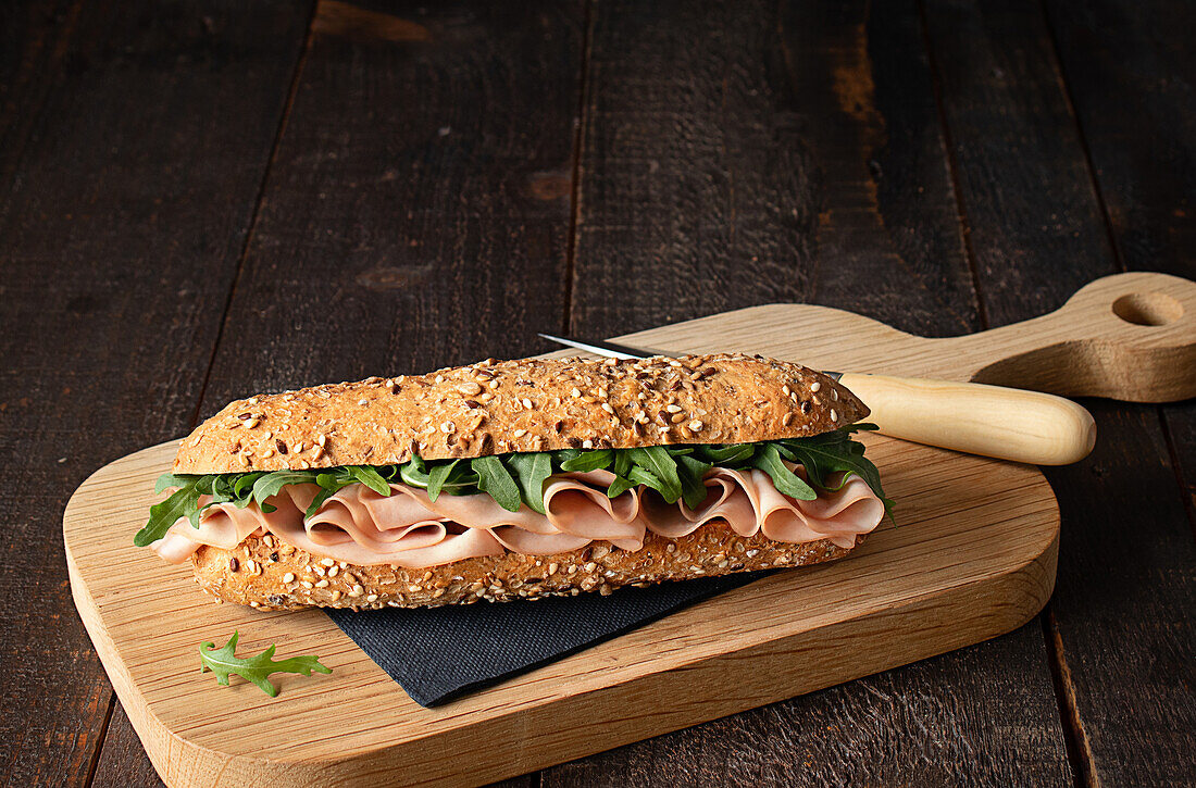 Mortadella-Schinken-Sandwich mit Rucola-Blättern auf hölzernem Schneidebrett auf rustikalem Küchentisch