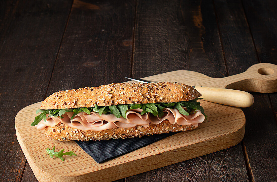 Mortadella ham sandwich with rocket leaves on wooden cutting board on rustic kitchen table