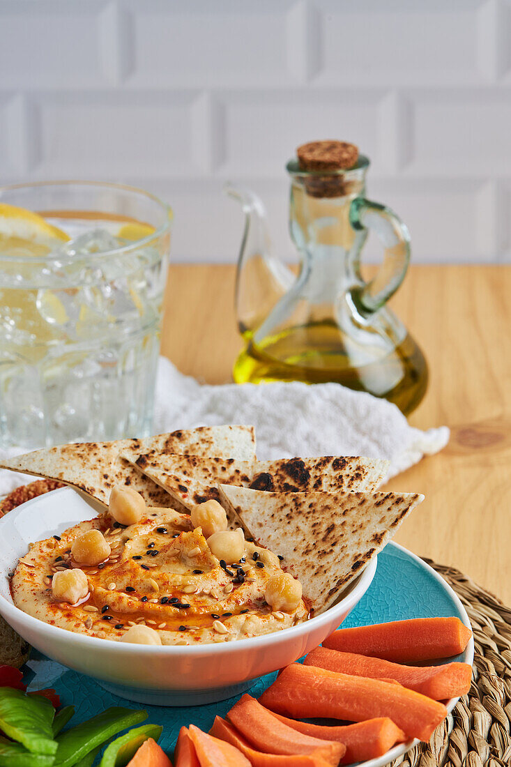 Schale mit Hummus und Tortilla-Chips, umgeben von verschiedenen Gemüsesorten und Croutons, serviert auf einer Weidenmatte neben Olivenöl und Limonade auf einem Holztisch von oben