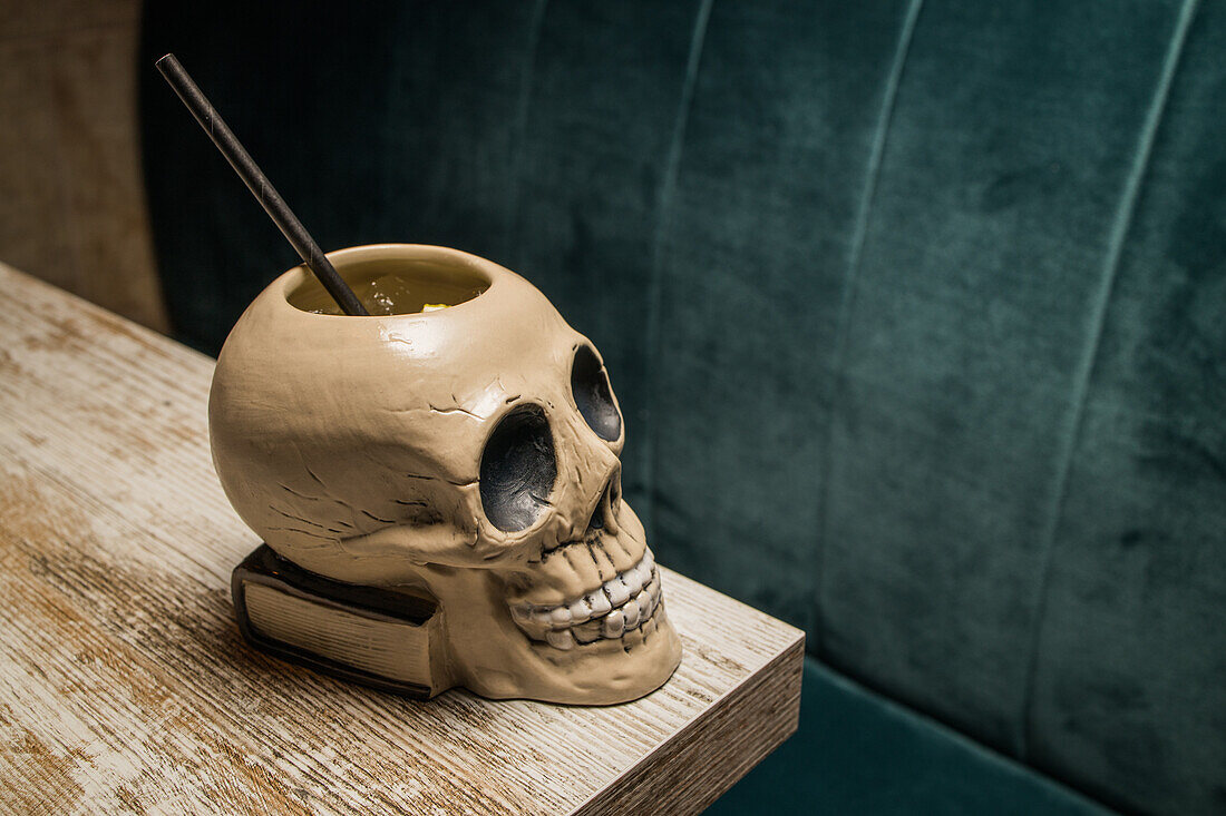 Ceramic polynesian tiki cup skull shaped with straw placed on wooden table on blurred background