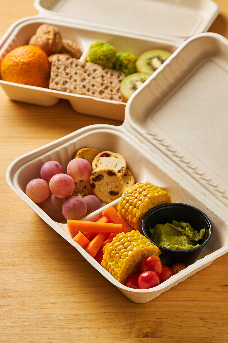 Von oben von Lunch-Boxen mit gesunden Lebensmitteln einschließlich Cracker Karotten-Sticks Trauben Kirschtomaten mit Kiwi Brokkoli Walnuss und Mandarine auf rosa Hintergrund