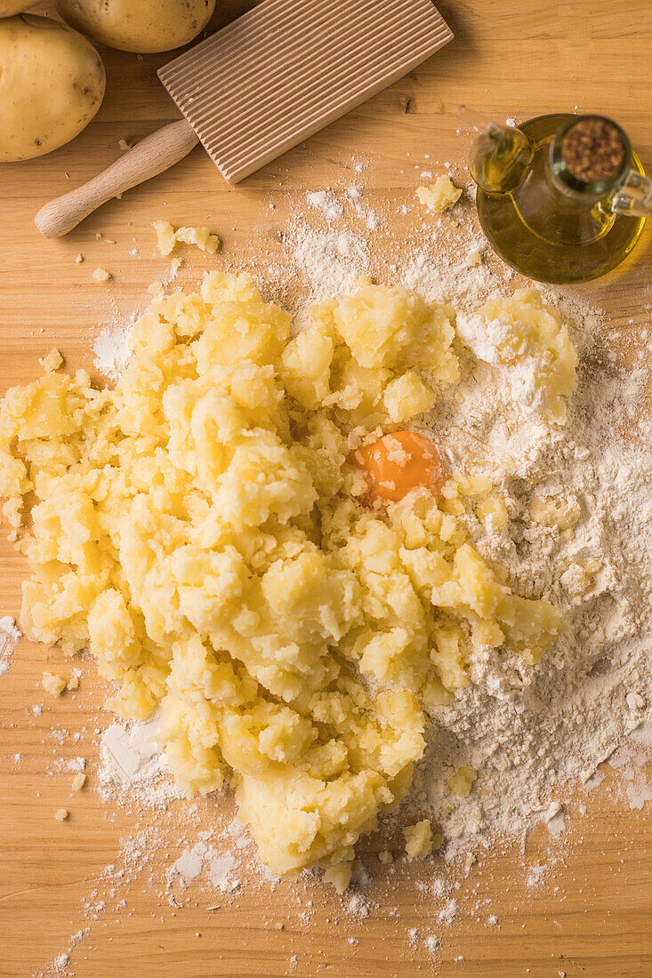 Blick von oben auf Eigelb in einem Haufen Kartoffelpüree und Weizenmehl in der Nähe von Öl und geripptem Brett bei der Zubereitung von Gnocchi auf dem Tisch