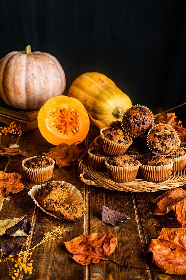 Composition made of appetizing muffins with chocolate chips placed on wooden table among scattered autumn leaves and pumpkins on dark background