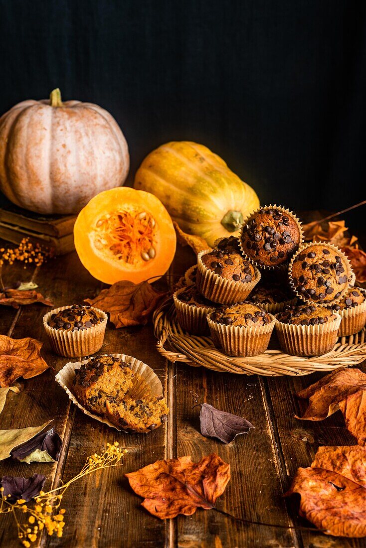 Komposition aus appetitlichen Muffins mit Schokoladenstückchen auf Holztisch zwischen verstreuten Herbstblättern und Kürbissen auf dunklem Hintergrund