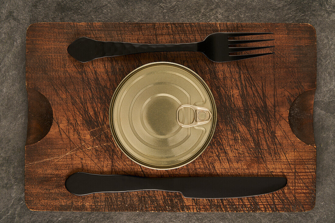 From above scratched chopping board with fork and sealed can with preserved food on rustic lumber table