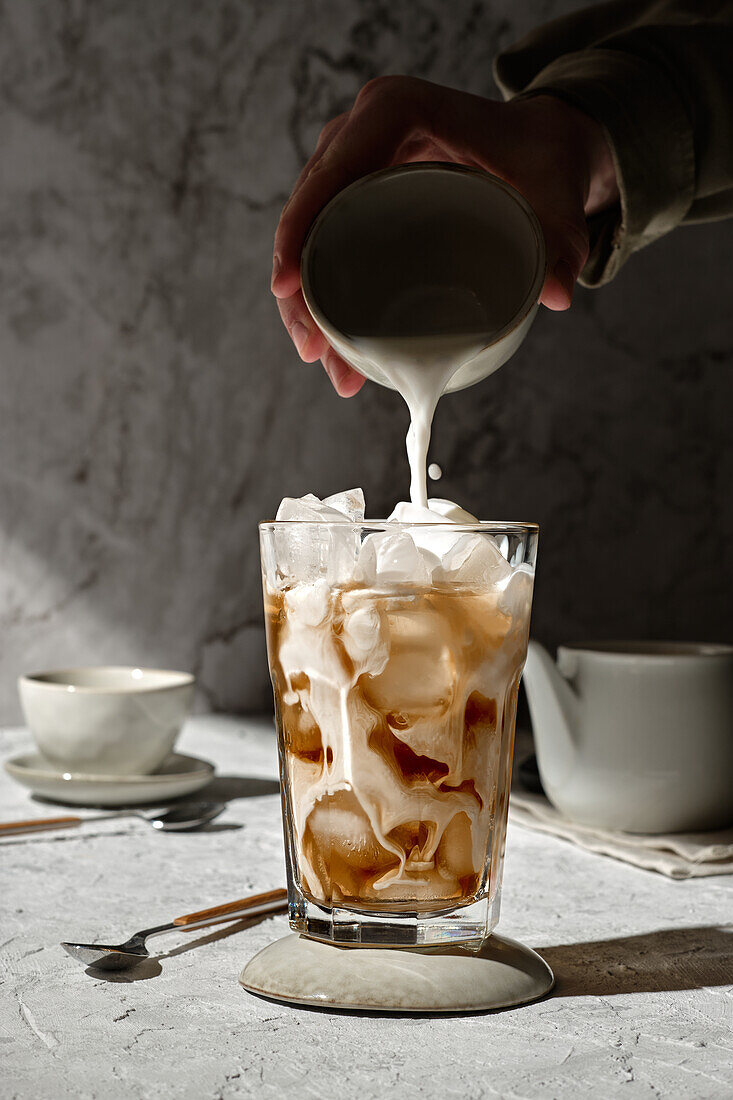 Anonyme Person gießt Milch aus einer Tasse in ein Glas mit Eiskaffee, das auf einem Tisch im Sonnenlicht steht