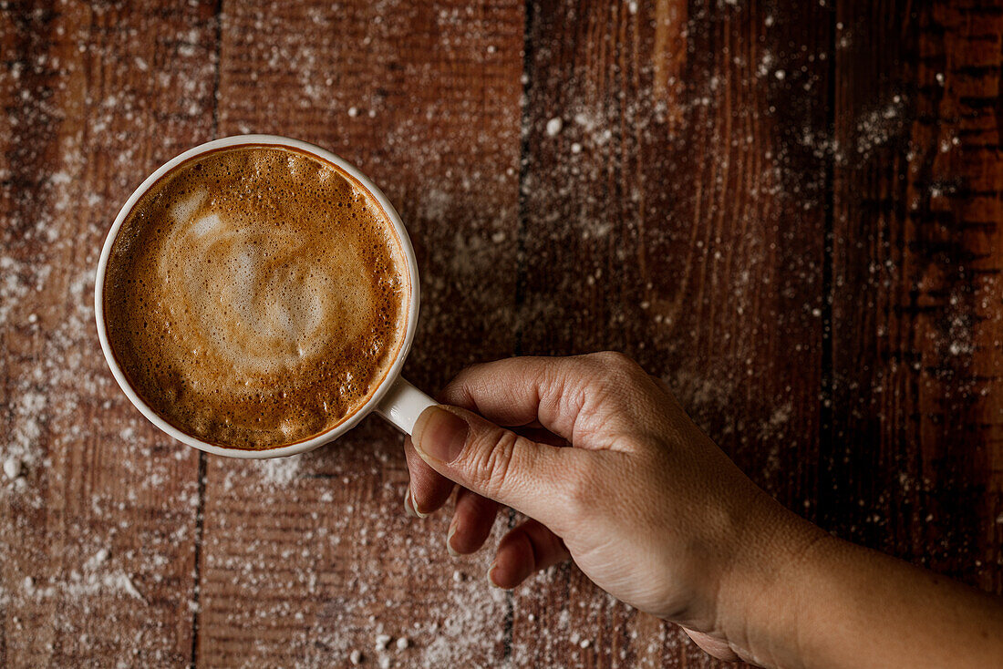 Top view of crop anonymous person with cup of aromatic coffee with foam on wooden table