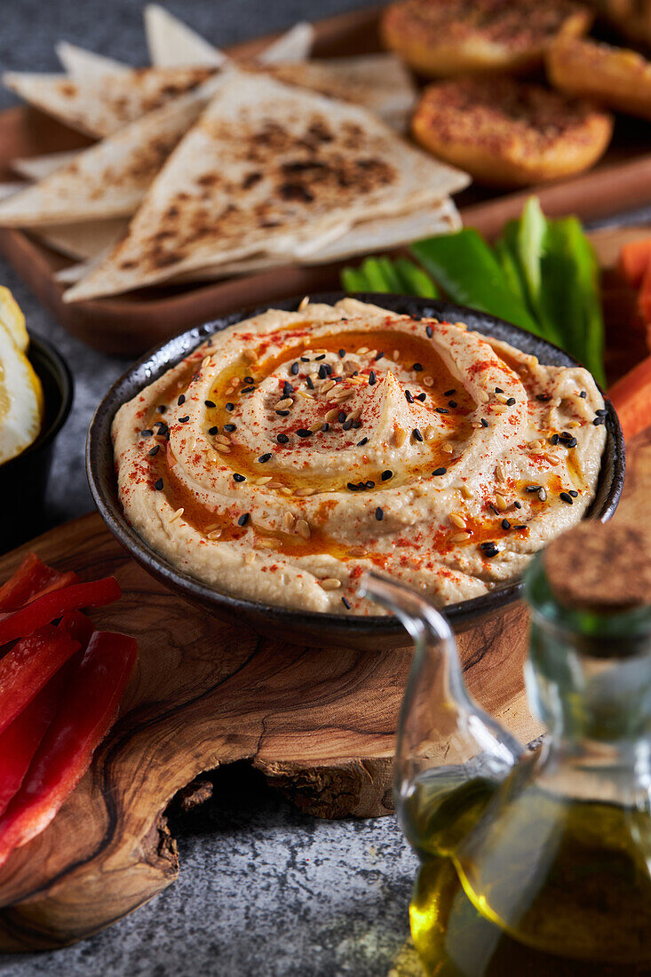 Schale mit leckerem Hummus auf Holzplatte mit verschiedenen Gemüsesorten neben Zitronenscheiben und Ölkrug vor Tortilla-Chips und Croutons