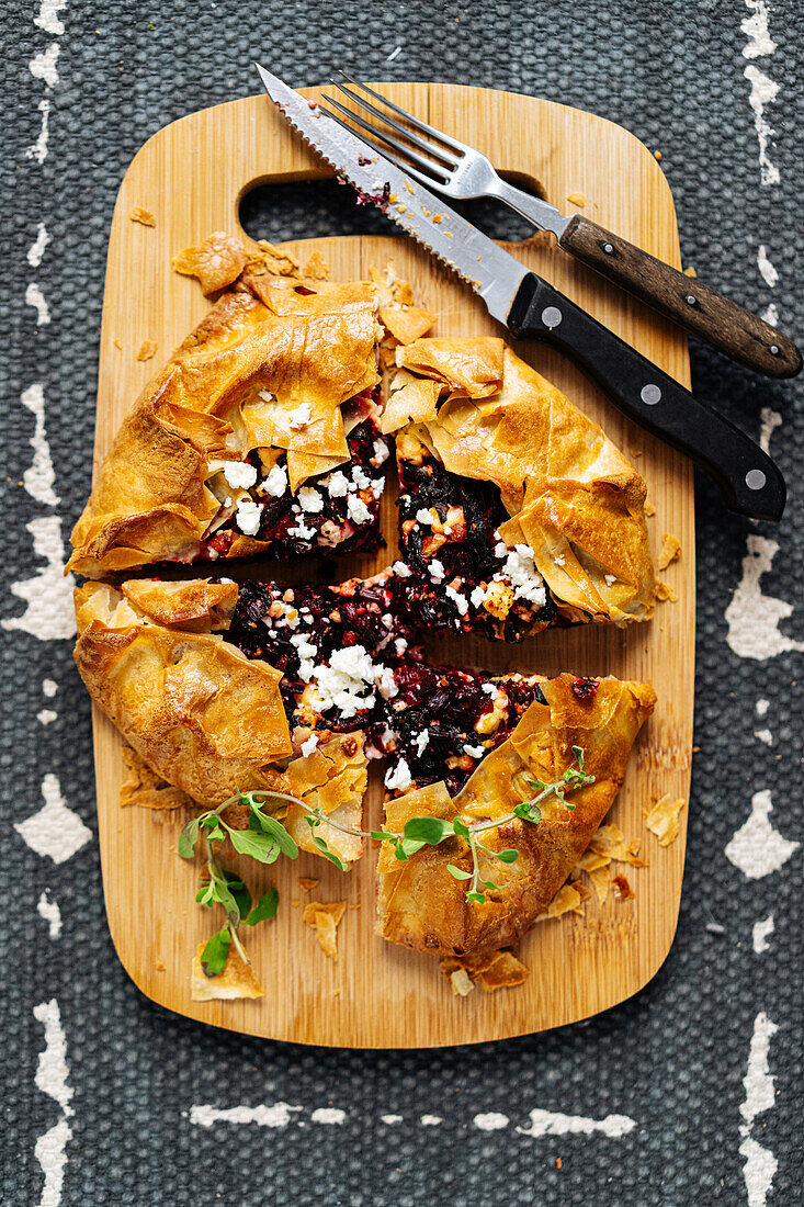 Top view of baked cut pie with beetroot and feta cheese on wooden cutting board with knife on gray background in kitchen