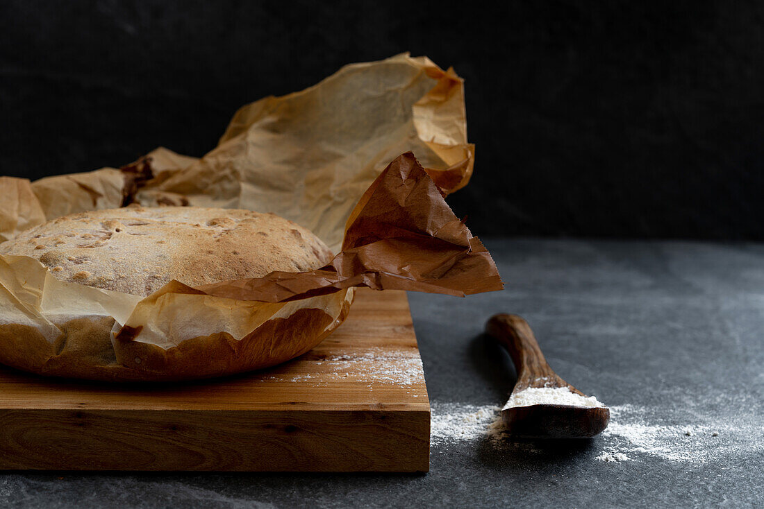 Composition with freshly baked rustic sourdough round bread loaf on parchment paper placed on wooden board with spoon and wheat flour