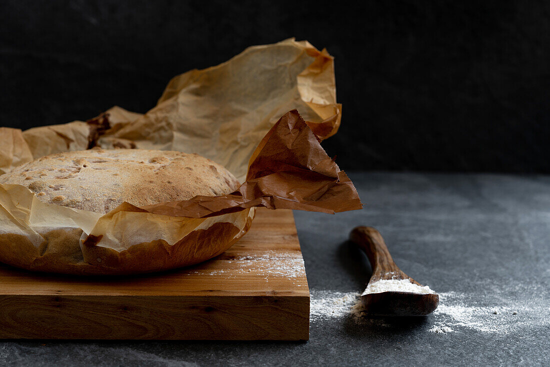 Komposition mit frisch gebackenem, rustikalem, rundem Sauerteigbrot auf Pergamentpapier auf einem Holzbrett mit Löffel und Weizenmehl