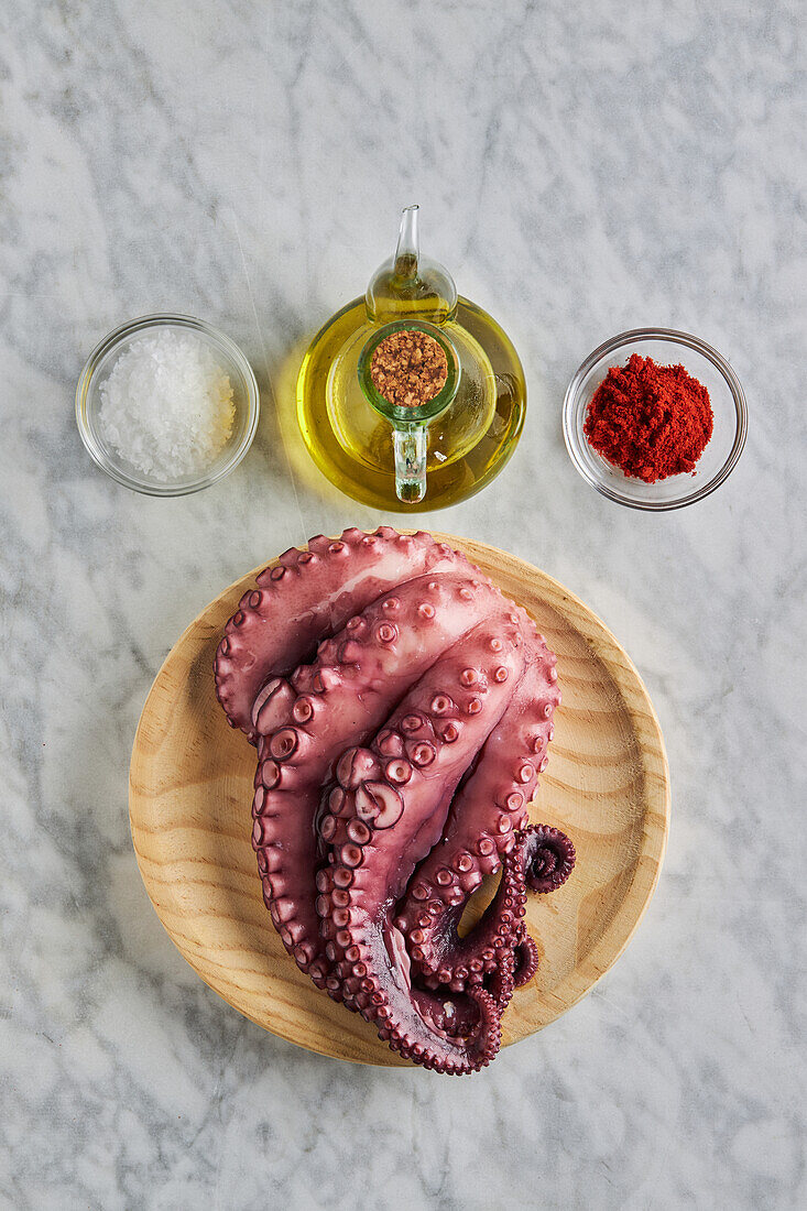 Draufsicht auf frischen rohen Oktopus auf einem runden Holzteller neben Gewürzen und Öl