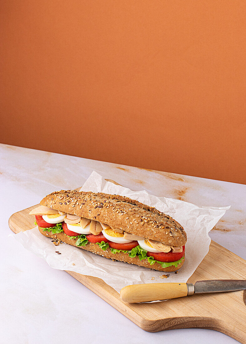 Appetitliches Sandwich mit hartgekochtem Ei, frischem Salat, Thunfisch und Tomaten, serviert auf Backpapier auf einem Holzbrett mit buntem Hintergrund