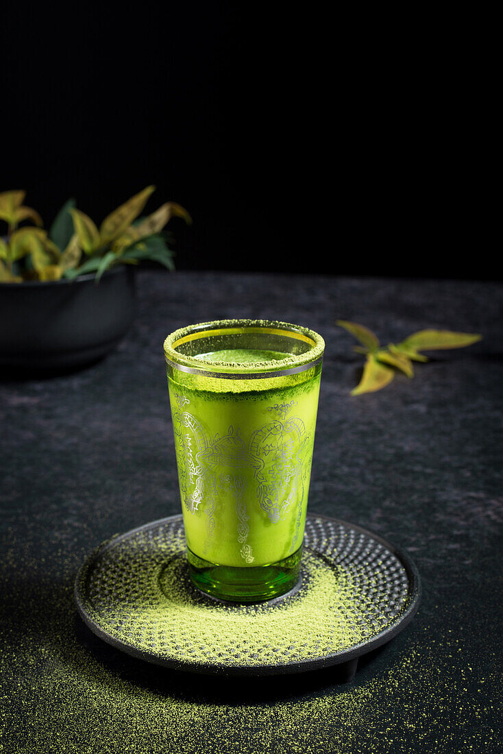 Still life composition with traditional oriental matcha tea served in glass cup with metal ornamental decor on table with ceramic bowls and fresh green leaves against black background