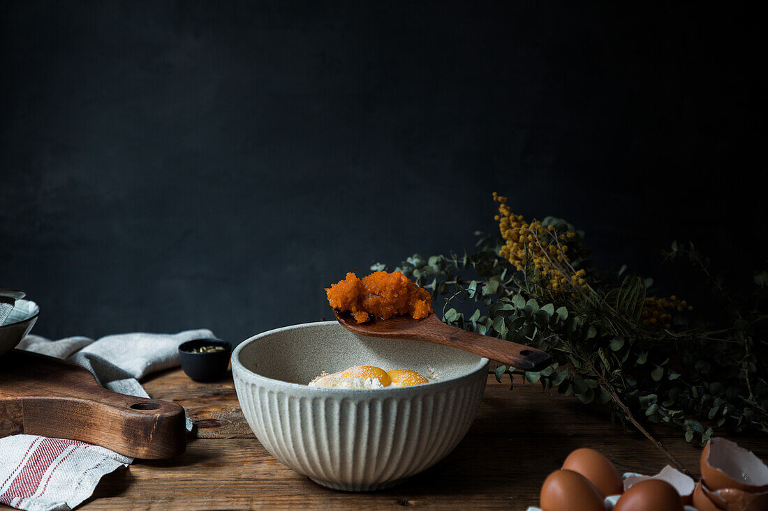 Bowl with eggs, flour and wooden spoon with pumpkin puree on wooden table during pastry preparation on dark background