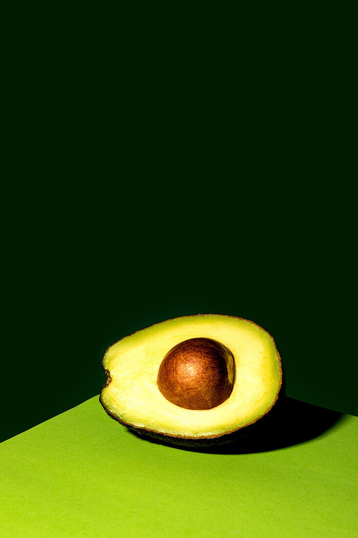 Eine halbe reife Avocado mit Samen liegt in einer Tischecke vor einem dunkelgrünen Hintergrund