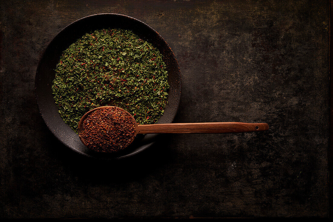 Overhead composition with spoon with ground sun dried tomatoes placed near bowl with green herbal spice on black background