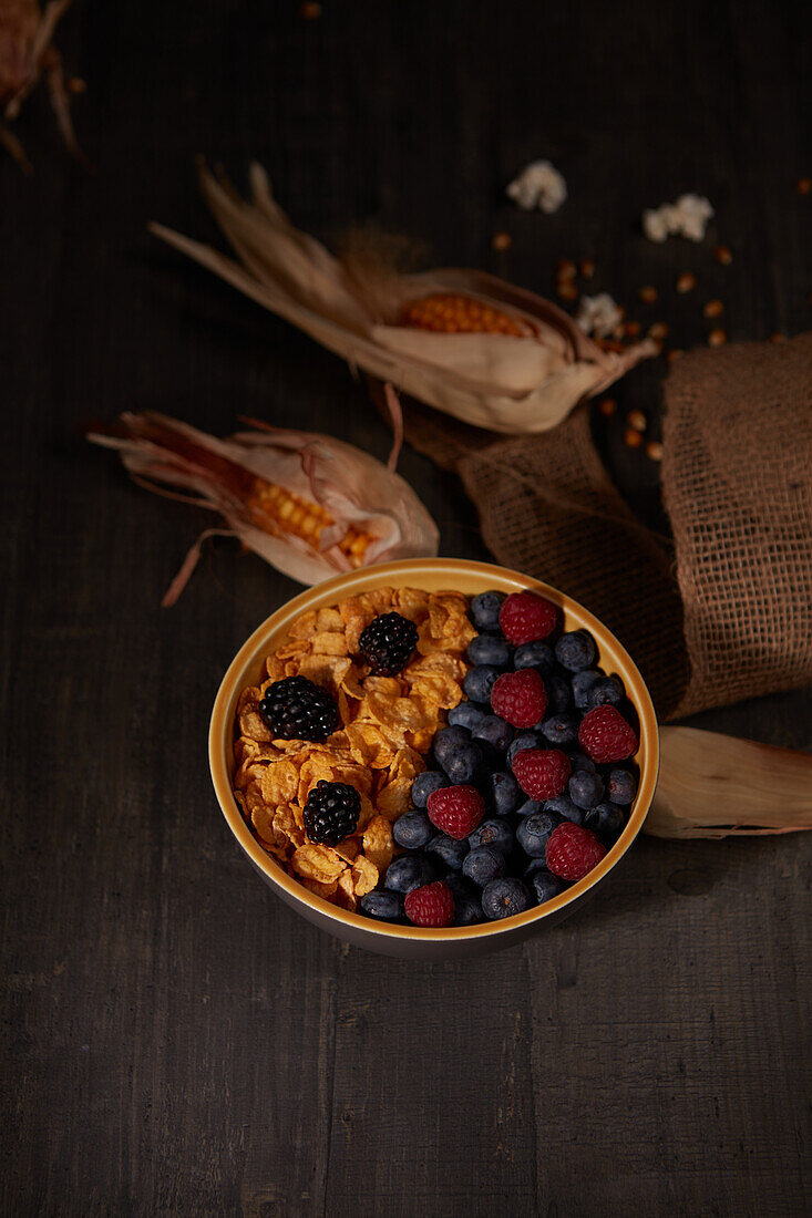 From above tasty cornflakes with fresh forest berries served in bowl near burlap fabric during breakfast in dark room