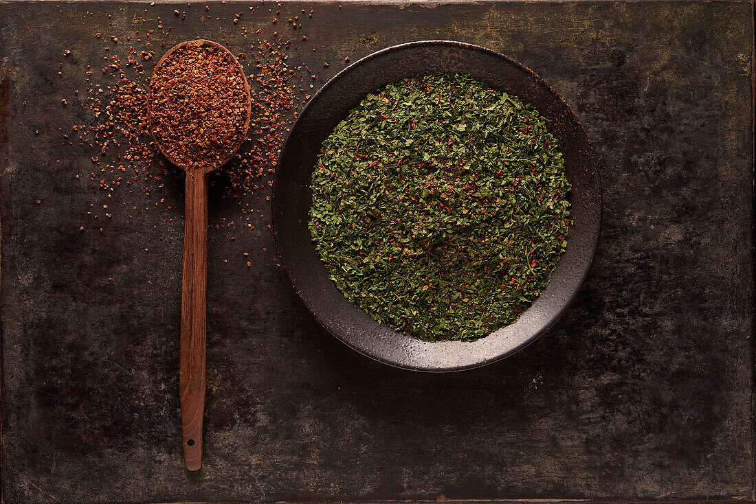 Overhead composition with spoon with ground sun dried tomatoes placed near bowl with green herbal spice on black background