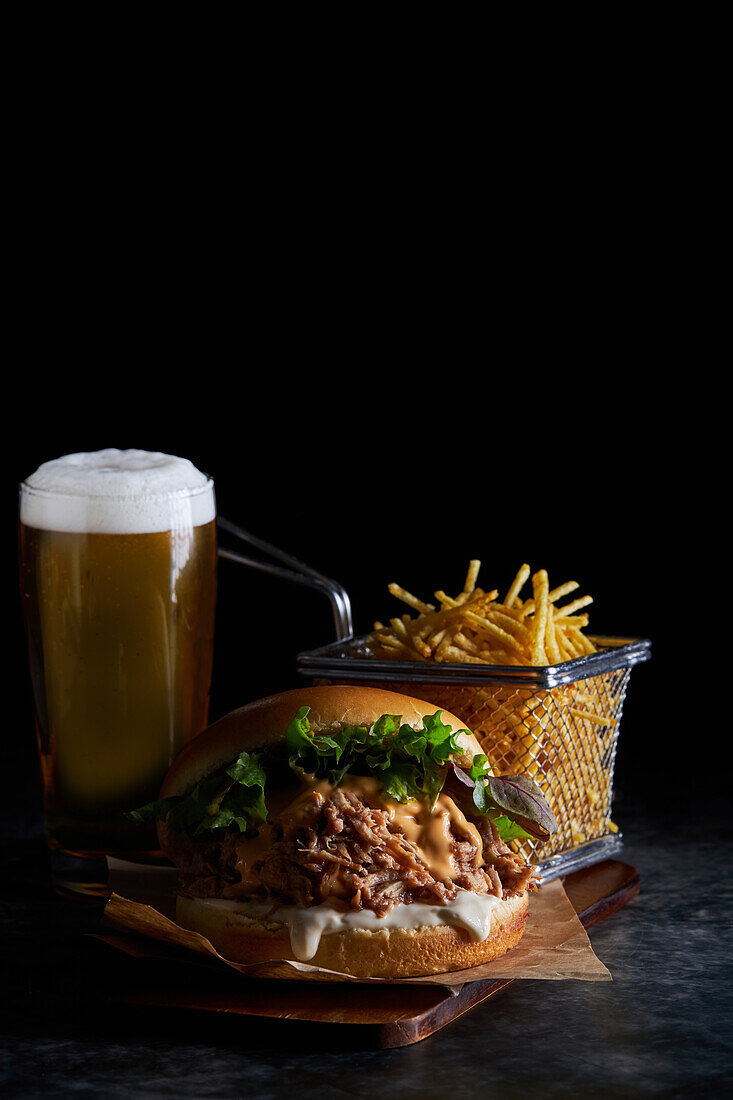 Appetizing burger with pulled pork with barbecue sauce cheddar cheese and lettuce mix served with French fries and glass of cold beer on wooden board