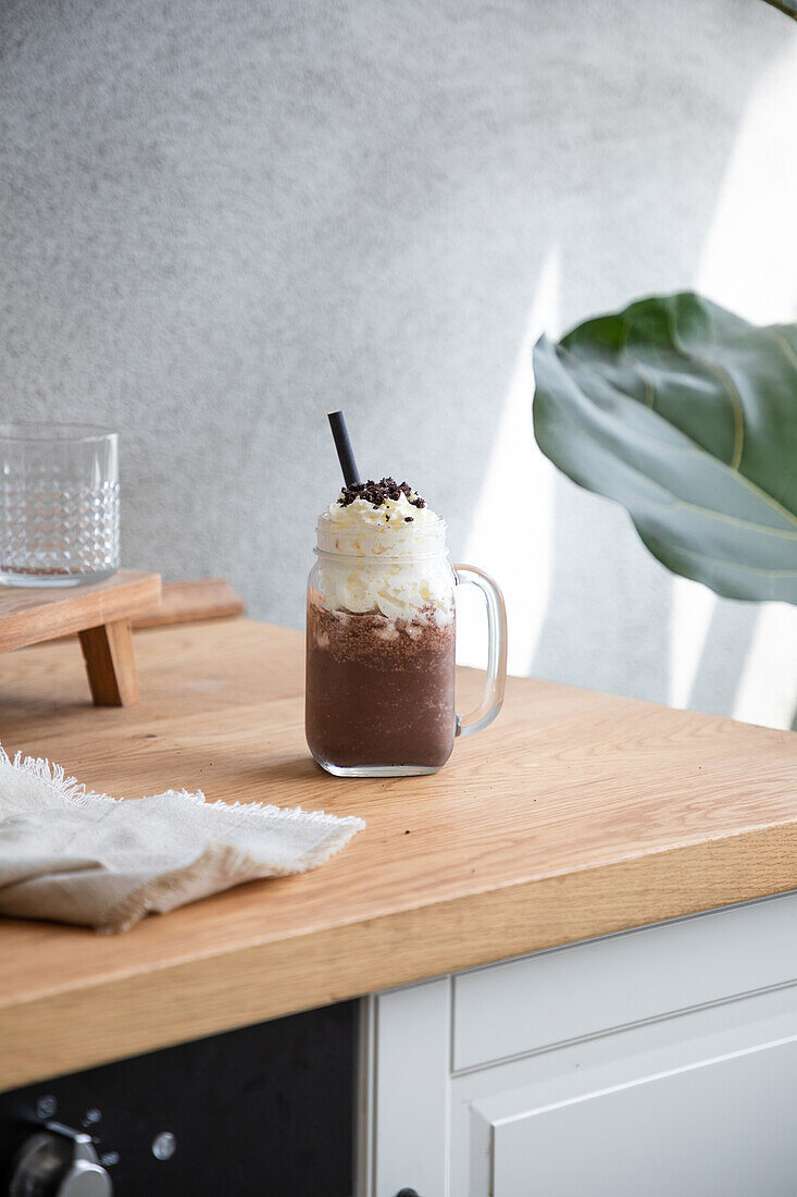 Glass jar with chocolate frappe topped with sweet whipped cream served with straw placed on wooden table in light room