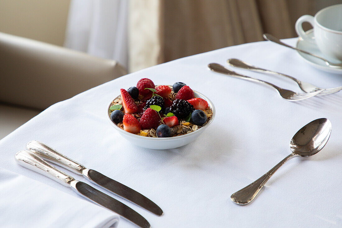 Von oben Schüssel mit gesundem Müsli mit frischen Beeren auf weißem Tisch mit Silberbesteck und Kaffeetasse beim Frühstück serviert