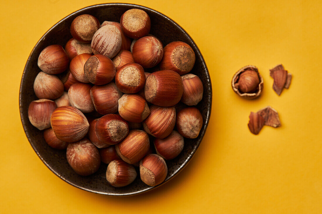 Top view of fresh unpeeled hazelnuts placed in bowl near cracked nut on yellow background