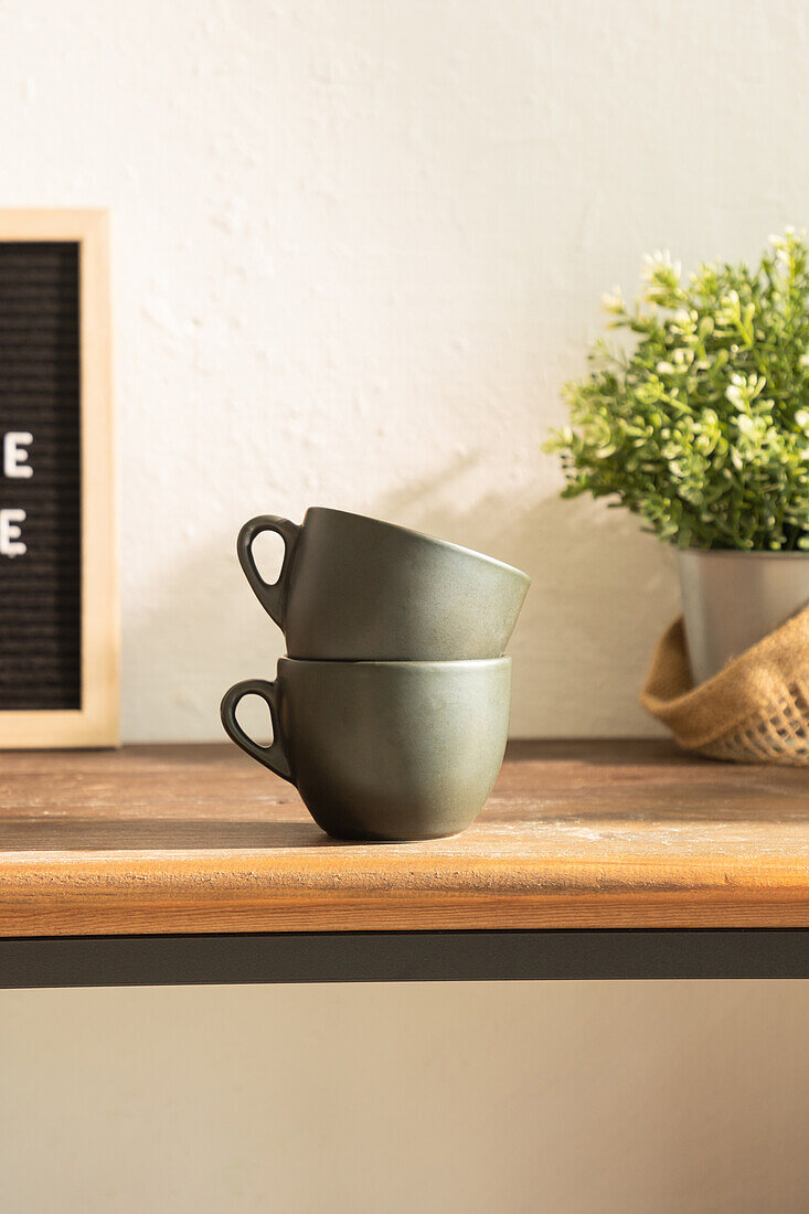 Mugs placed on each other on wooden counter with black signboard and green potted plant in light kitchen at home