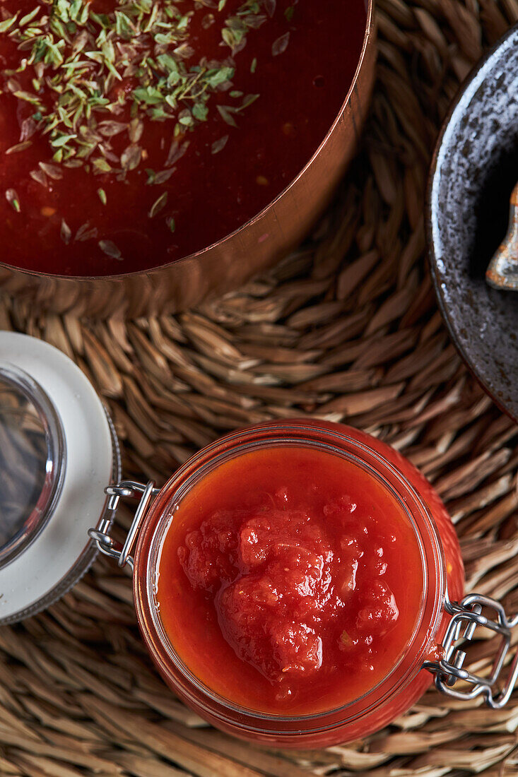 Draufsicht auf Pfanne und Behälter mit köstlicher hausgemachter Tomatensauce mit Oregano bestreut auf gewebter Matte auf Holztisch in Küche