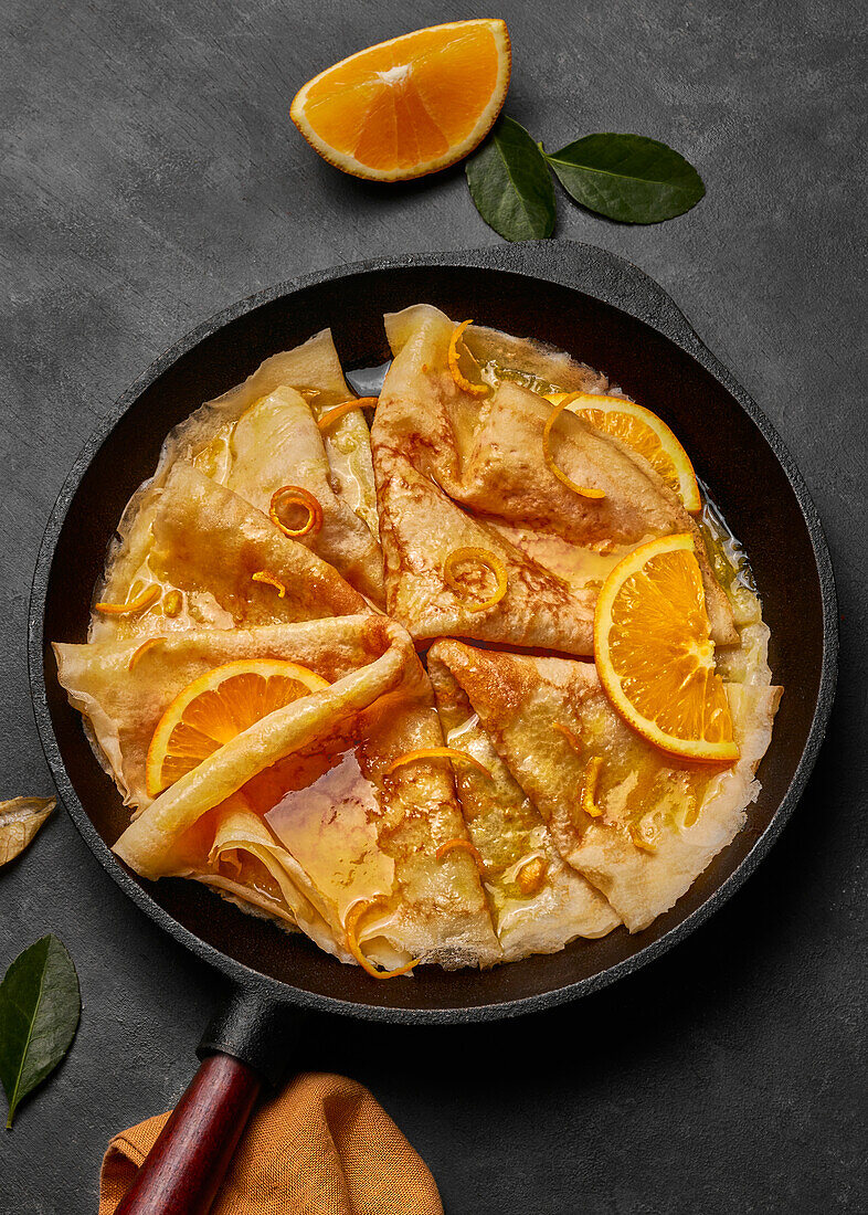 From above homemade crepes suzette with orange liqueur and orange slices served on plate with knife and fork on concrete surface