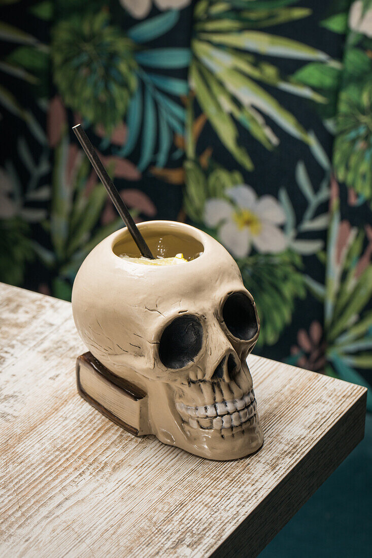 Keramische polynesische Tiki-Tasse in Form eines Totenkopfes mit Strohhalm auf einem Holztisch vor unscharfem Hintergrund