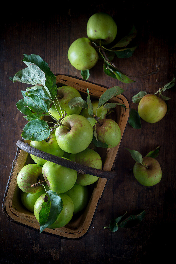 Blick von oben auf einen Haufen ganzer kleiner grüner Äpfel mit Blättern in einem Korb auf einer Holzfläche