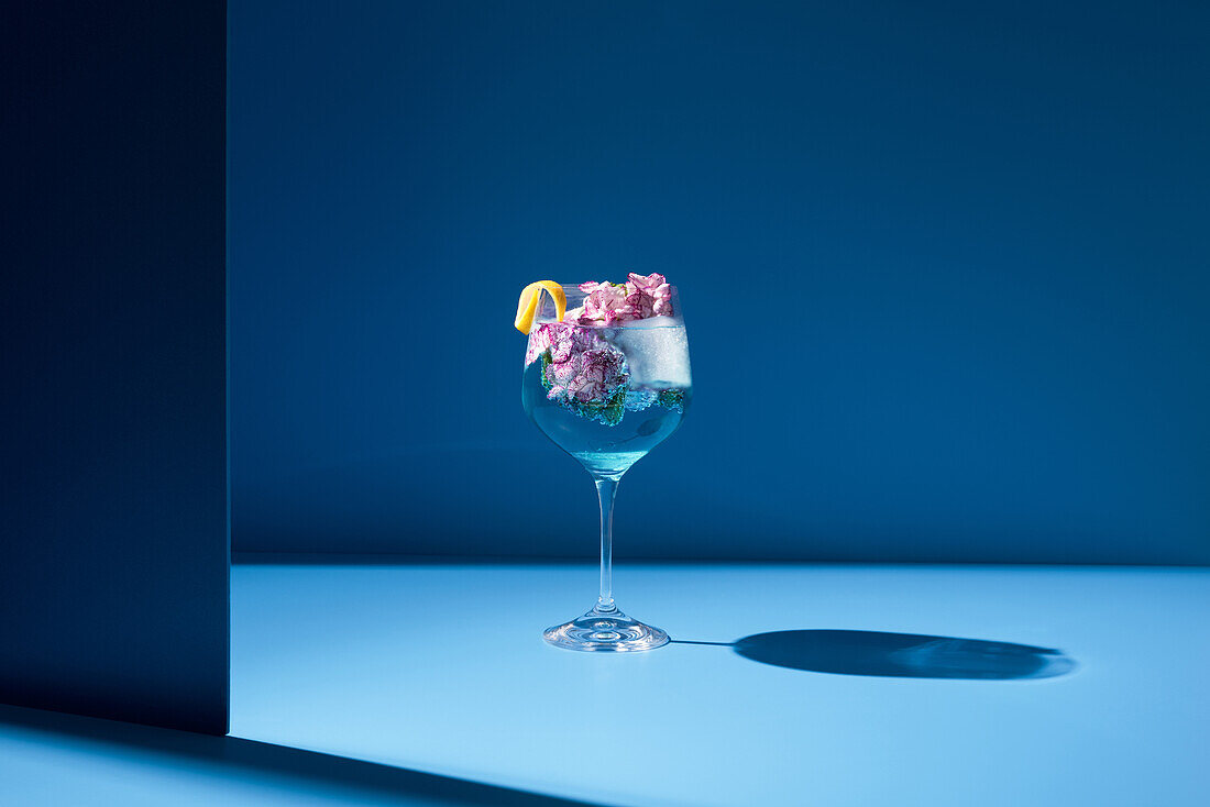 Transparentes Glas mit frischem Cocktail, Minzblättern und Blumen vor blauem Hintergrund auf dem Tisch