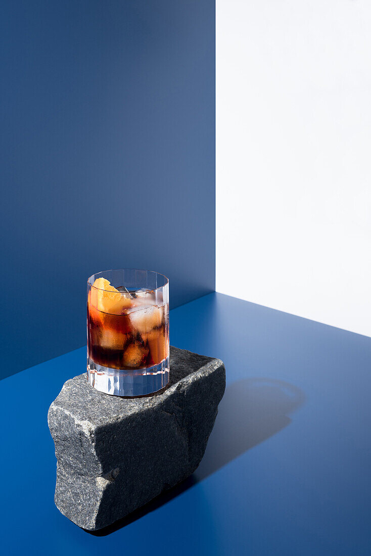Glas alkoholischer Cocktail mit rotem Wermut, serviert mit Orangenscheiben auf einem Stein in einem hellen Studio mit blauer und weißer Wand mit Schatten
