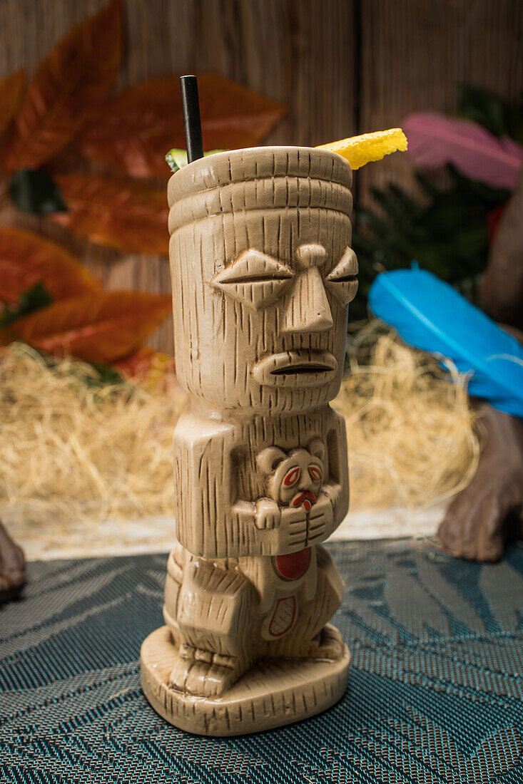Traditional sculptural tiki cup of alcohol drink with straw placed on rug against wooden fence colorful leaves and dry grass