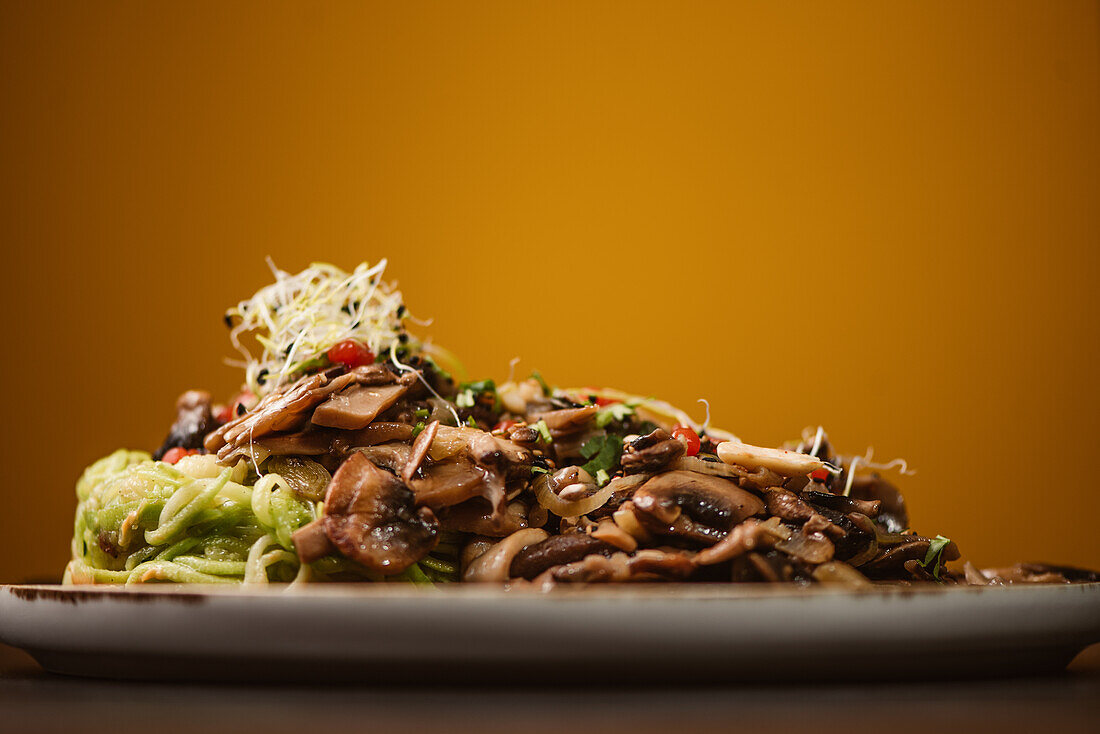 Low angle of yummy vegan dish with zucchini spaghetti and sauteed mushroom slices covered with red berries and alfalfa sprouts on yellow background