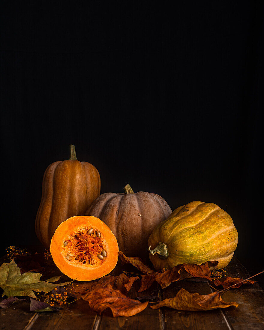 Composition of ripe orange pumpkins and dry autumn maple leaves placed on wooden table near black wall