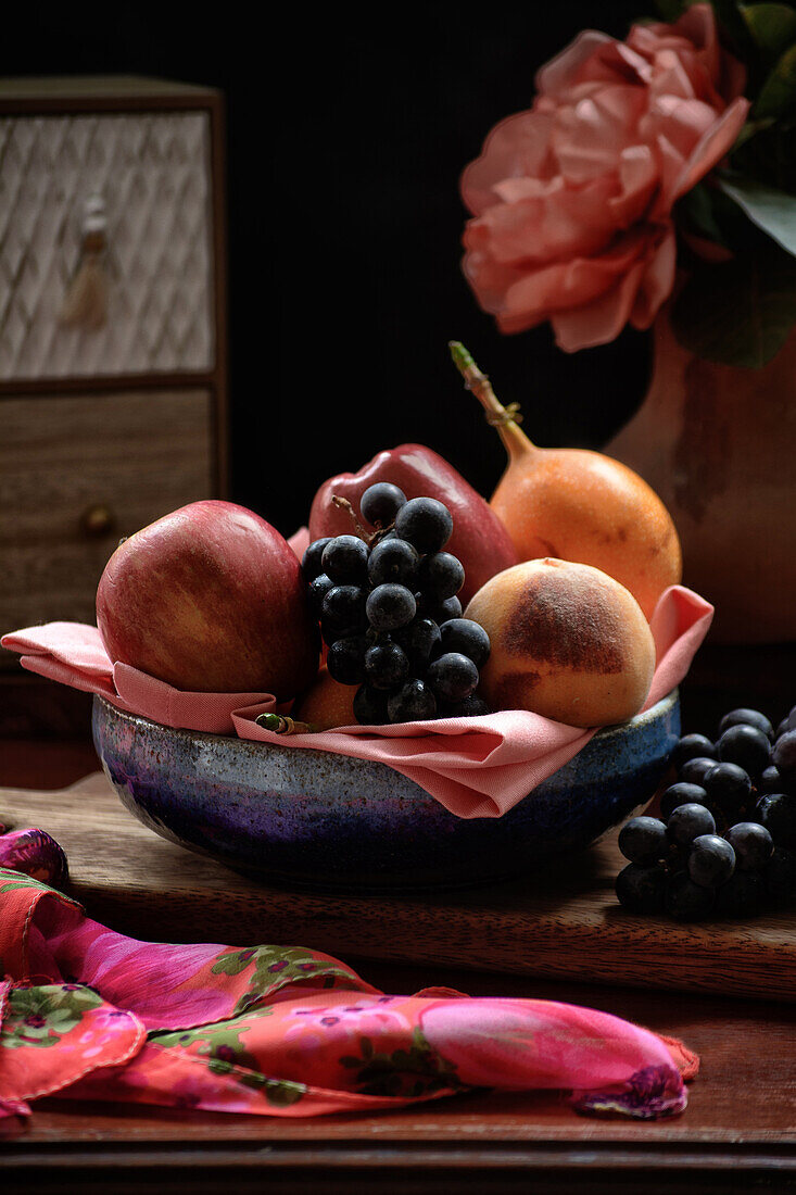 Frische Äpfel und Pfirsiche mit Weintrauben und Granadilla in einer Schale auf einem Tisch neben einer roten Pfingstrose