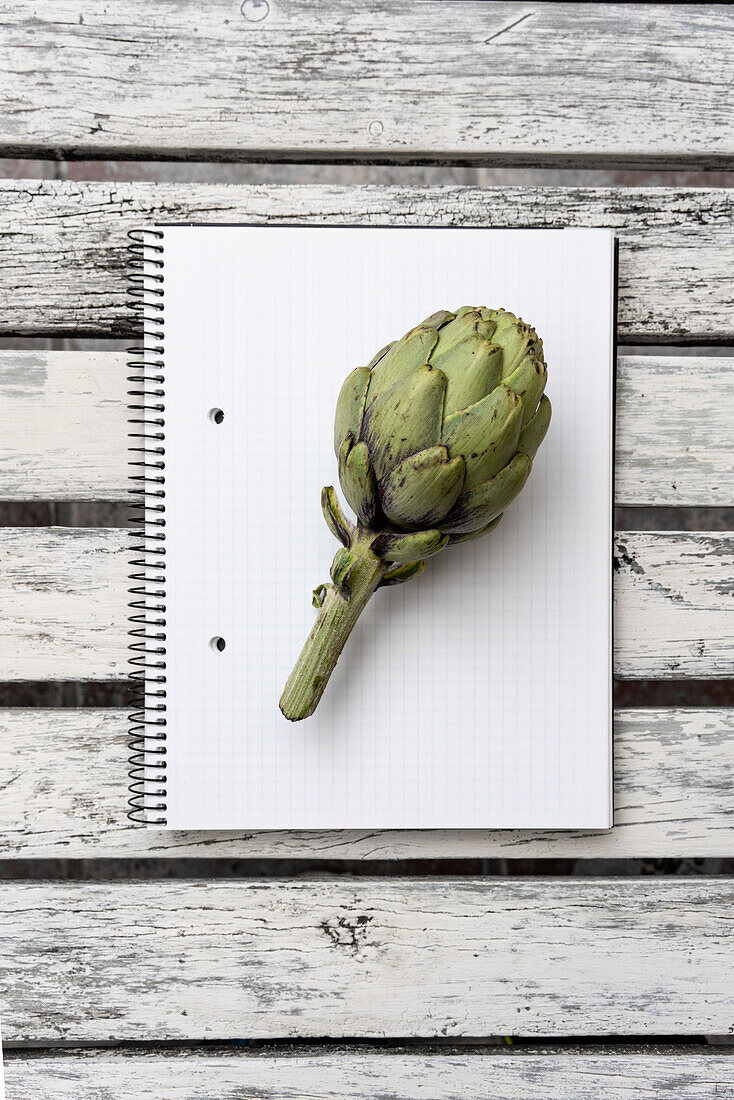 Top view of green artichoke placed on opened notepad with blank pages on wooden table