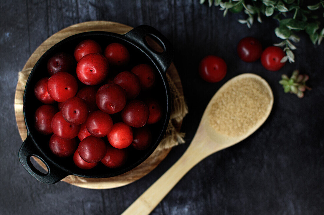 Schale mit frischen süßen Pflaumen von oben auf schwarzem Tisch mit Holzlöffel mit Zucker serviert