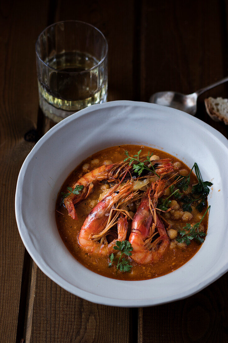 From above of delicious spicy stew with chickpeas and big shrimps decorated with cilantro leaves served in white soup plate on wooden table with glass of drink and spoon