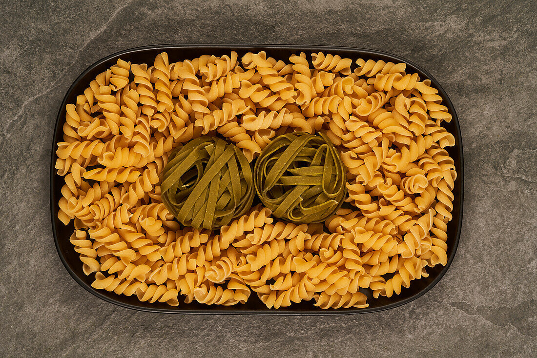 Top view of tray filled with raw fusilli and tagliatelle pasta and placed on gray table
