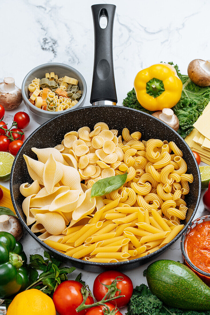 From above of frying pan with various uncooked pasta surrounded with assorted raw vegetables on marble table in light kitchen