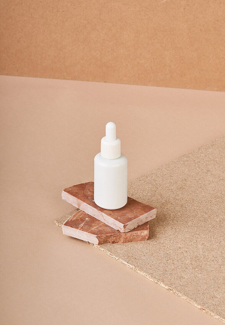 Kleine weiße Flasche mit kosmetischem Serum auf gestapelten braunen Marmorsteinen auf Stoff vor beigem Hintergrund