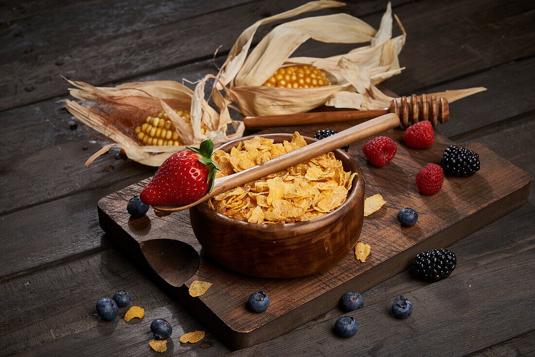 Bowl of tasty sweet cornflakes with spoon on wooden chopping board placed on table with corn cobs and berries in light kitchen