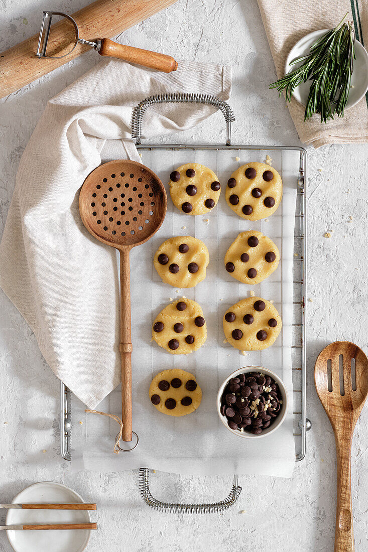 Top view of freshly baked sweet cookies with chocolate chips on metal grid placed on table with various kitchen tools and green rosemary branches