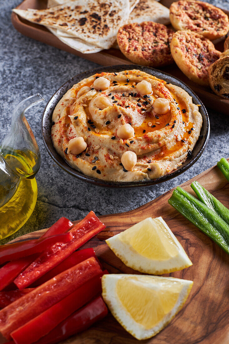 High angle of bowl with tasty hummus placed near wood slab with assorted vegetables near slices of lemon and pitcher of oil against chips and croutons