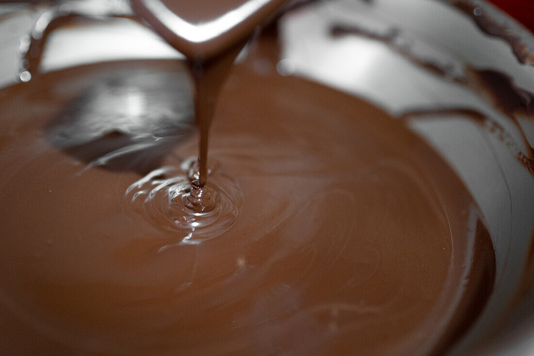 Anonymer Angestellter mit Handschuhen benutzt einen Metallspatel, um flüssige Schokolade auf dem Tisch zu verteilen, während er ein Dessert in einer Konditorei zubereitet